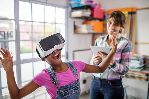 Surprised schoolgirl wearing virtual reality headset by teacher. Female student is learning through virtual reality in classroom. They are in elementary school.