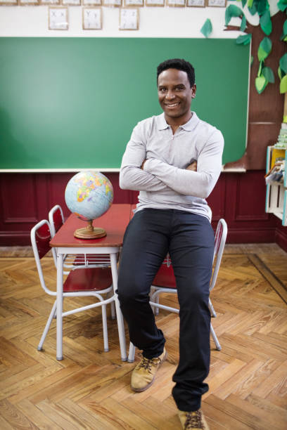 Portrait of smiling male teacher sitting arms crossed on table. Full length of confident man is with globe in classroom. He is wearing casuals in elementary school.
