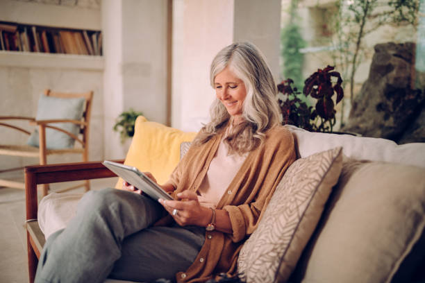 улыбаясь старшая женщина с седыми волосами с помощью планшета в домашних условиях - relaxation indoors reading one person стоковые фото и изображения