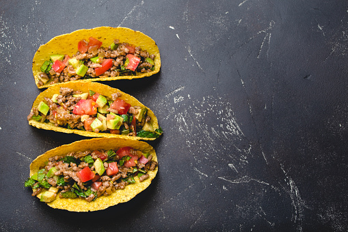 Top view of Mexican tacos with meat, tomato salsa, avocado, herbs in yellow corn tortilla on black rustic stone background with copy space. Traditional Mexican dish for lunch or dinner