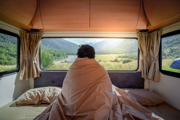 Young Asian man staying in the blanket looking at mountain scenery through the window in camper van in the morning. Road trip in summer of South Island, New Zealand. Young Asian man staying in the blanket looking at mountain scenery through the window in camper van in the morning. Road trip in summer of South Island, New Zealand. camper trailer photos stock pictures, royalty-free photos & images