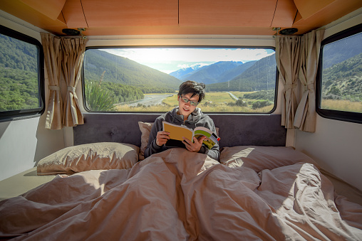 Young Asian man staying in the blanket reading magazine book in camper van with mountain scenery through the window. Road trip in summer of South Island, New Zealand.