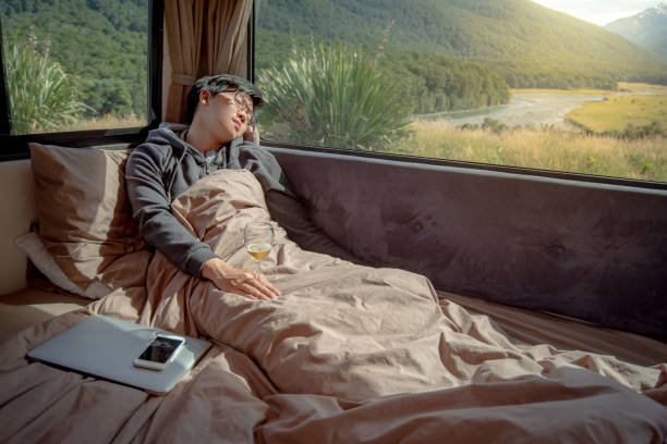 istock Young Asian man hangover after drinking beer in camper van with mountain scenic view through the window, laptop computer and smartphone on the bed. digital nomad on road trip concept 997023558