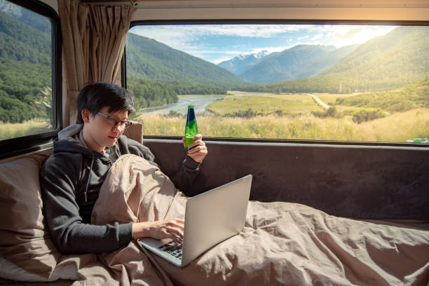 istock Young Asian man drinking beer and working with laptop computer on the bed in camper van with snow mountain scenic view through the window, digital nomad on road trip concept 997023560