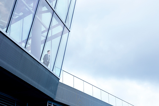 A businessman stands with his hands in his pockets inside a large office building and looks through the window into the distance. The overcast sky provides ample room for copy and text.