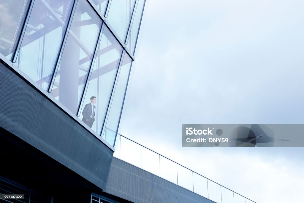 Businessman Stands Inside Building And Looks Out Through Window A businessman stands with his hands in his pockets inside a large office building and looks through the window into the distance. The overcast sky provides ample room for copy and text. Building Exterior Stock Photo
