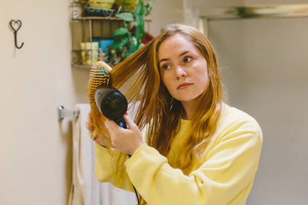 Young Adult Blow Drying and Brushing Hair Teenage female using a mirror in her home bathroom. brushing hair stock pictures, royalty-free photos & images