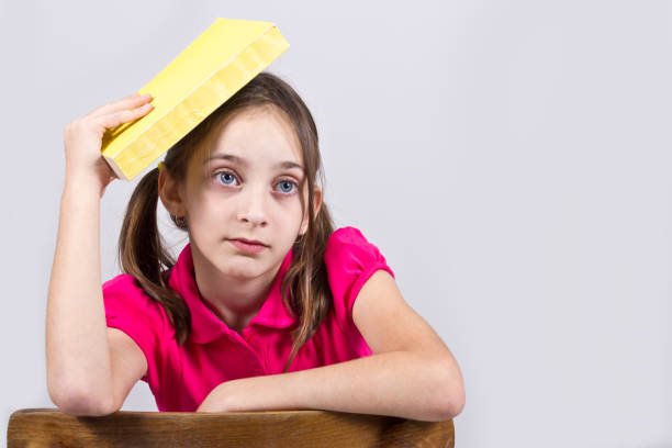A young girl  hitting herself in the head with a book and looking aside .Learning concept, studio shoot isolated on grey
