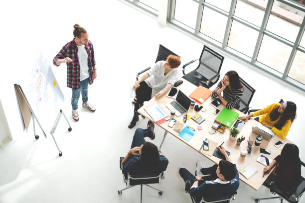 istock Young caucasian creative man standing and making presentation at modern office happy talking and brainstorming with team. Casual multiethnic people business meeting concept with wide angle top view. 997323904