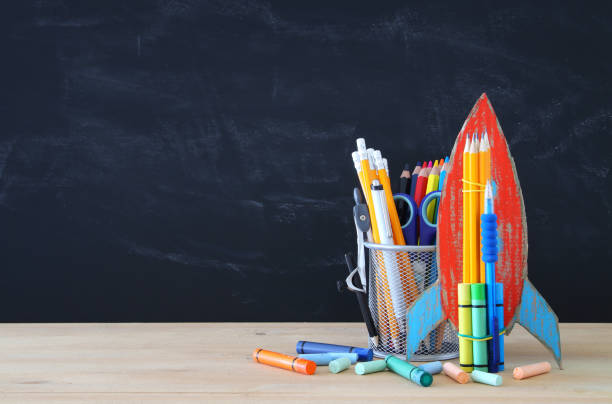 istock Back to school concept. cardboard rocket and pencils over open book in front of classroom blackboard. 997341442