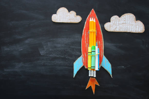 istock Back to school concept. Top view image of handmade cardboard rocket and clouds with pencils over classroom blackboard background 997341580