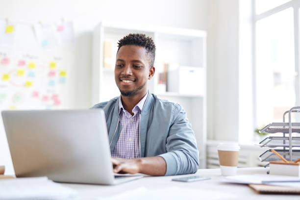 Positive young African-American male copywriter in casual jacket typing on laptop and sitting at table while working on article in office Positive African copywriter in office copywriter photos stock pictures, royalty-free photos & images