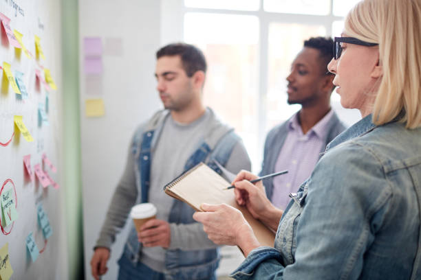 istock Pensive office lady asking question and making notes during brainstorming with colleagues, they standing at board full of adhesive notes 997398026