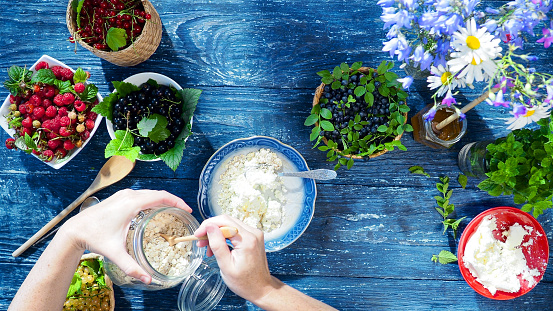 Healthy summer breakfast with fresh berries, farmer's cheese and oatmeal on the blue wooden table. Human hands are putting oats into the plate. Overhead view