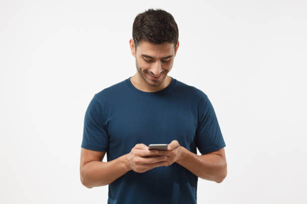 closeup photo of casually-dressed european male standing isolated on gray background looking attentively at screen of cellphone, browsing web pages and smiling nicely while chatting with friends - attentively imagens e fotografias de stock