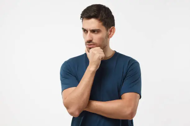 Photo of Skeptical and dissatisfied young man in blue t-shirt touch his chin with hand. Doubt concept