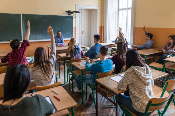 Large group of teenage students with raised arms in the classroom. Rear view of high school students raising hands to answer teacher's question during the class. teenage high school girl raising hand during class stock pictures, royalty-free photos & images