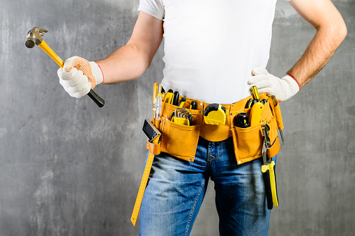 unidentified handyman standing with a tool belt with construction tools and holding a hammer against grey background. DIY tools and manual work concept