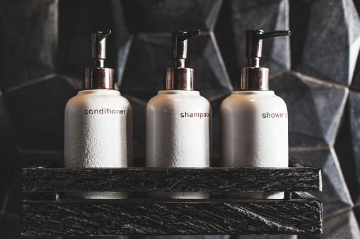 Three white metal bottles of shampoo, conditioner, shower gel on wooden shelf  in modern hotel bathroom with textured black wall