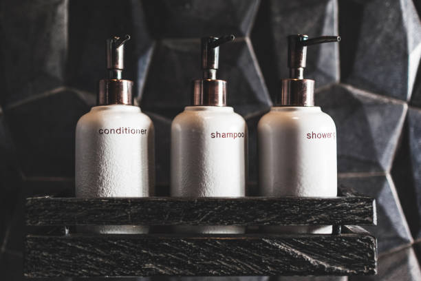 Three white metal bottles of shampoo, conditioner, shower gel on wooden shelf in modern hotel bathroom with textured black wall stock photo