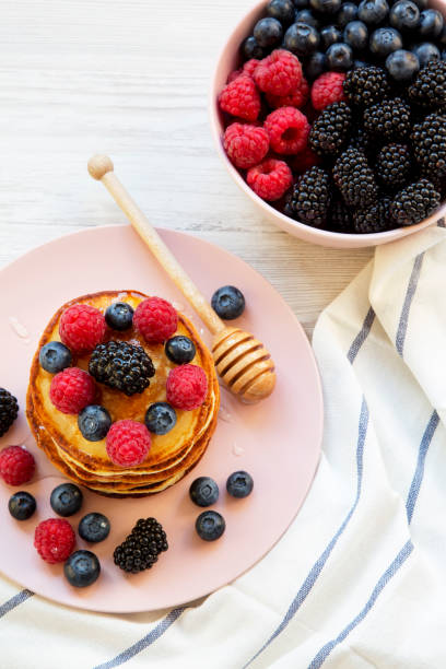 Pancakes with berries and honey on a pink plate, bowl of berries, high angle. Close-up.