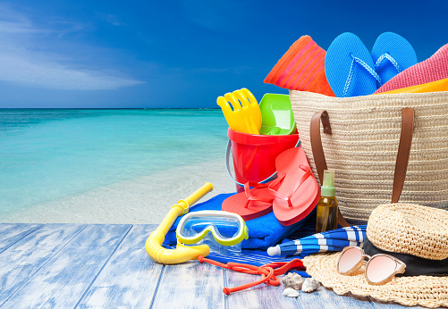 Front view of multi colored beach accessories shot on blue plank with turquoise colored beach at background. Accessories included in the composition are a beach umbrella, beach towels, a pair of flip-flops, scuba mask with snorkel, suntan lotion, beach bag, sun hat and a sunglasses. The accessories are grouped at the right of the frame leaving useful copy space for text and/or logo at the center-left. Beach, summer or vacations concept. DSRL outdoors photo taken with Canon EOS 5D Mk II and Canon EF 24-105mm f/4L IS USM Wide Angle Zoom Lens
