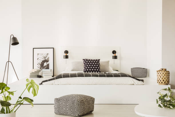 istock Patterned pouf in front of white bed in bright bedroom interior with plant, lamp and poster. Real photo 997536574