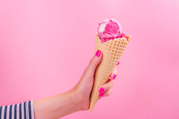 Woman's hand holding ice cream waffle cone with berry and vanilla ice cream scoop on pink background