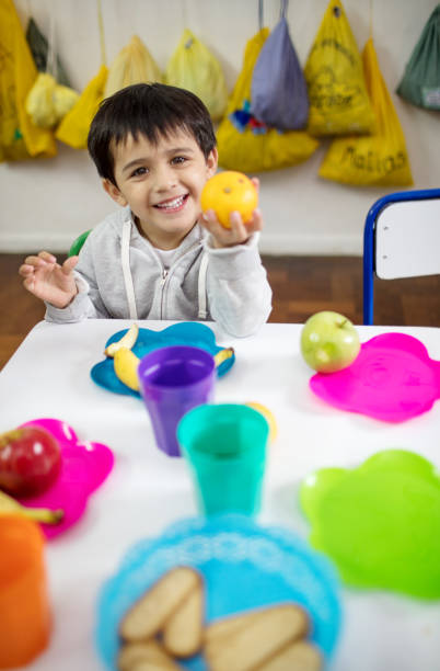 Preschool kid eating fruits in classroom Portrait of happy little boy showing a orange while sitting at desk during lunch break. Preschool kid eating fruits in classroom. food elementary student healthy eating schoolboy stock pictures, royalty-free photos & images