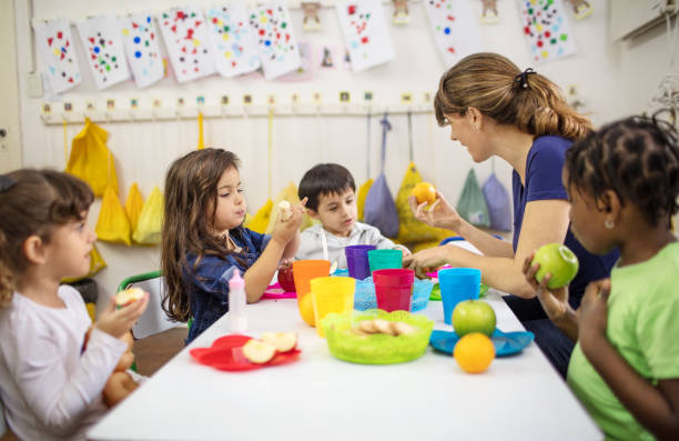Teacher explaining fruits to students in classroom Preschool teacher explaining students about fruits. Woman sitting with kids at desk during lunch. preschool building photos stock pictures, royalty-free photos & images