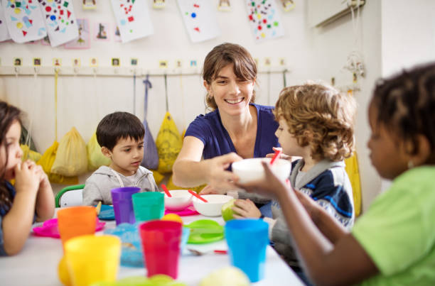 Teacher with preschool kids at desk during lunch Happy female teacher giving bowl to girl while sitting with students in classroom. Woman with preschool kids at desk during lunch. preschool building stock pictures, royalty-free photos & images