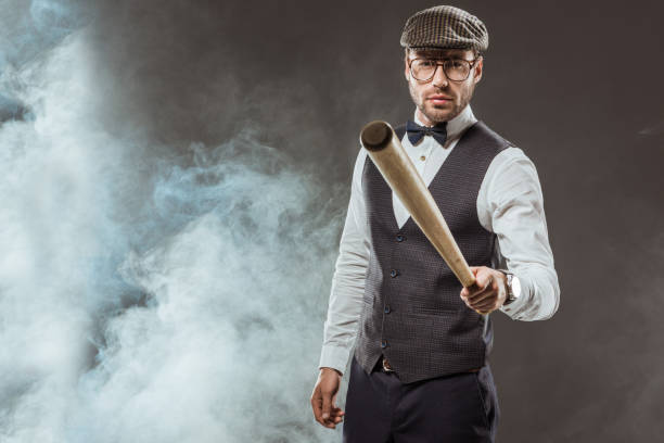 istock serious man in bow tie and cap holding baseball bat while standing in smoke 997579798
