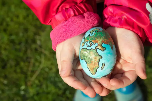 Photo of Child holding an egg with Planet Earth painted on it on a sunny day outdoors