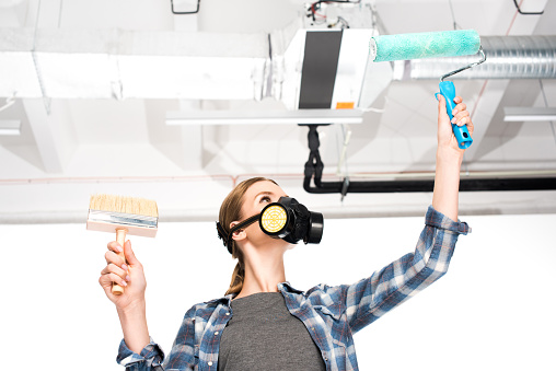 low angle view of woman in respirator holding paint roller and paint brush with pipes on behind