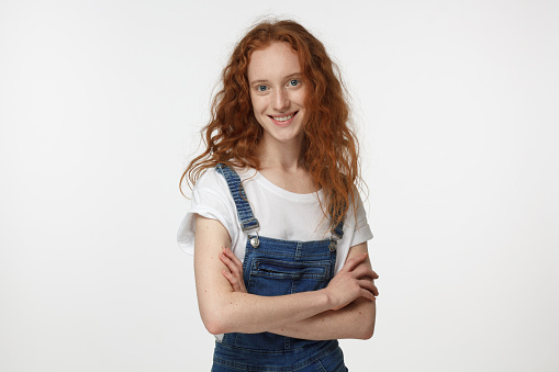 Portrait of smiling beautiful redhead girl in jean jumpsuit and white t-shirt standing with crossed arms isolated on gray background