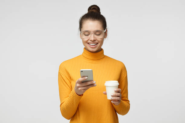 Indoor picture of young good-looking European woman isolated on gray background holding phone in hand, looking to screen smiling while browsing or reading message Indoor picture of young good-looking European woman isolated on gray background holding phone in hand, looking to screen smiling while browsing or reading message nerd teenager stock pictures, royalty-free photos & images