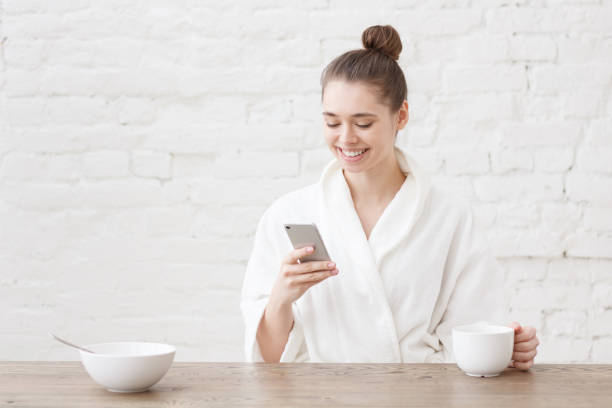 istock Young woman sitting at white loft kitchen, after bath, looking at her smartphone, having delicious breakfast, checking messages, holding coffee cup in one hand 997677904