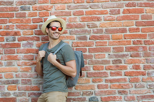 Horizontal image of young Caucasian man pictured on left side of red brick background watching what is happening in street walking in urban surrounding with backpack and hat on one summer day