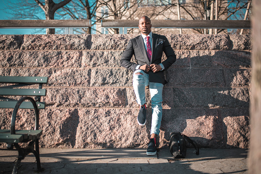 Shot of black businessman on the go leaning on rocky wall in New York City, USA. Blue sky with sun in the back.