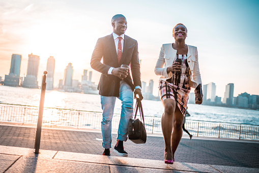 Shot of black businessman and African-American business woman on the go holding coffee cup and digital tablet and smiling in New York City, USA. City and skyscrapers in the back.