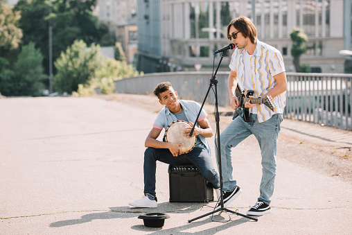 Young and happy male street musicians playing guitar and djembe in city