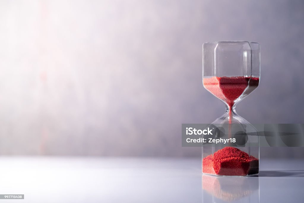 Red sand running through the shape of modern hourglass on white table.Time passing and running out of time. Urgency countdown timer for business deadline concept with copy space Hourglass Stock Photo