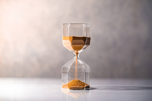 istock Brown sand running through the shape of modern hourglass on white table.Time passing and running out of time. Urgency countdown timer for business deadline concept 997715518