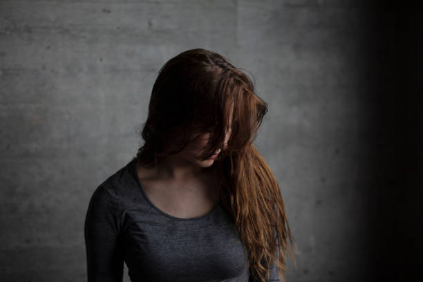 Female model posing behind a grey concrete wall and looking downwards. Medium shot.