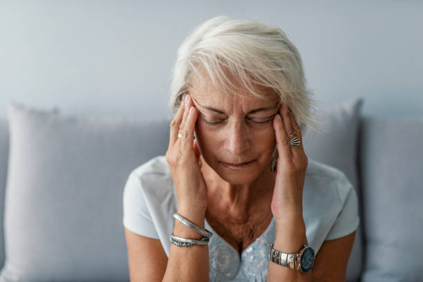 Senior woman having headache and touching her temples Senior woman having headache and touching her temples. Mature woman sitting on a white sofa in a home touching her head with her hands while having a headache pain and feeling unwell face down stock pictures, royalty-free photos & images