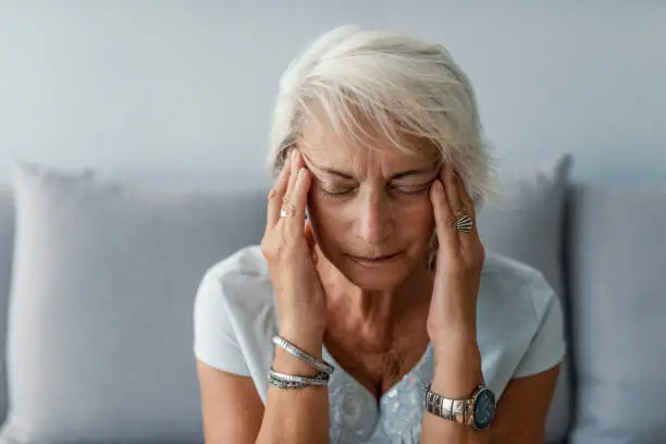 Senior woman having headache and touching her temples. Mature woman sitting on a white sofa in a home touching her head with her hands while having a headache pain and feeling unwell