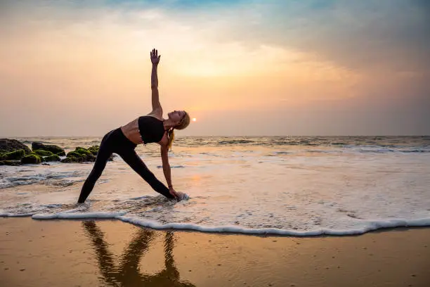 Middle age woman in black doing yoga on sand beach in India  standing asana Trikonasana (triangle pose) at sunset. Healthy lifestyle.