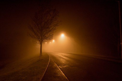 A road lined with orange glowing streetlights disappears into the gloom and fog