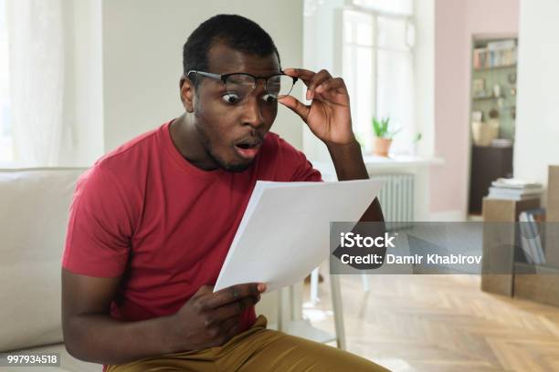 Photo libre de droit de Jeune Homme Afroaméricain Assis Sur Le Canapé Dans Un Appartement Moderne Avec La Bouche Ouverte Tenant La Facture Délectricité Avec Des Taux Élevés Déclenché Des Lunettes En Geste De Wow Ou De Surprise banque d'images et plus d'images libres de droit de Facture