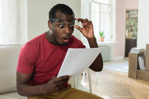 Photo of Young african american man sitting on couch in modern apartment with mouth open, holding utility bill with high rates, raised eyeglasses in wow or surprise gesture